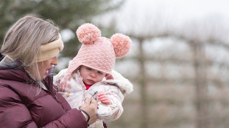 Woman carrying young child in a Garden, both wearing hats and gloves.